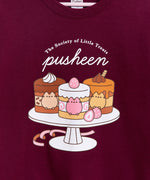 Close-up view of the cake graphic in the front center of the maroon sweatshirt. The three cakes sat on the cake stand are mocha, strawberry, and caramel. Inside the cakes are Pusheen the Cat figures that match their respective cake color.  