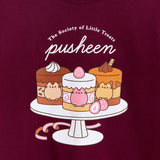 Close-up view of the cake graphic in the front center of the maroon sweatshirt. The three cakes sat on the cake stand are mocha, strawberry, and caramel. Inside the cakes are Pusheen the Cat figures that match their respective cake color.  