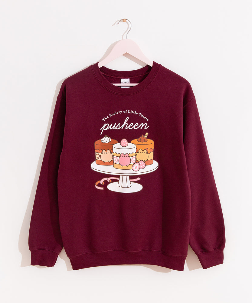 Pusheen Sweets Sweatshirt hangs on a light pink hanger in front of a wall. The maroon unisex sweatshirt features a large graphic on the front of the sweatshirt of three tiny cakes on a cake stand with the phrase “The Society of Little Treats Pusheen” above the cakes. 