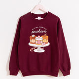 Pusheen Sweets Sweatshirt hangs on a light pink hanger in front of a wall. The maroon unisex sweatshirt features a large graphic on the front of the sweatshirt of three tiny cakes on a cake stand with the phrase “The Society of Little Treats Pusheen” above the cakes. 