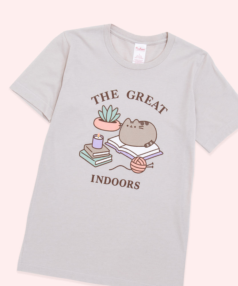 A light dusty beige graphic tee against a light-pink background. The center graphic features Pusheen sitting on an open book with the phrase, “The Great Indoors” in brown text above and below the Pusheen graphic. 