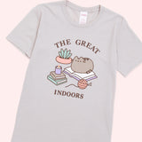 A light dusty beige graphic tee against a light-pink background. The center graphic features Pusheen sitting on an open book with the phrase, “The Great Indoors” in brown text above and below the Pusheen graphic. 