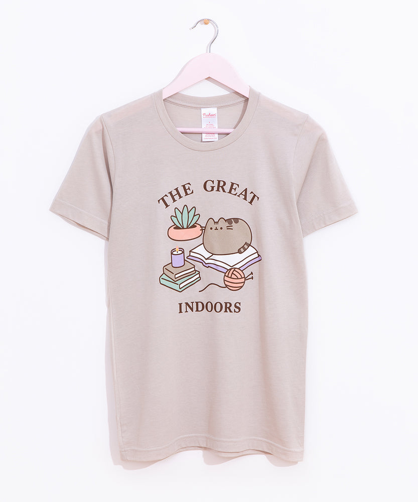 Pusheen The Great Indoors Unisex Tee hangs on a light pink hanger in front of a white background. The beige graphic tee has a large print in the center of the t-shirt. 