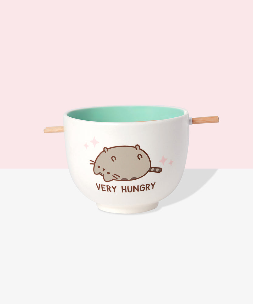 Front view of Pusheen Very Hungry Ramen Bowl and Chopstick Set. The chopsticks rest in a notch on the lip of the white bowl. A graphic of Pusheen belly up is on both sides of the bowl. Pink sparkles surround Pusheen and the phrase “Very Hungry” is printed in brown below the cat graphic. 