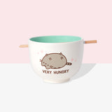 Front view of Pusheen Very Hungry Ramen Bowl and Chopstick Set. The chopsticks rest in a notch on the lip of the white bowl. A graphic of Pusheen belly up is on both sides of the bowl. Pink sparkles surround Pusheen and the phrase “Very Hungry” is printed in brown below the cat graphic. 