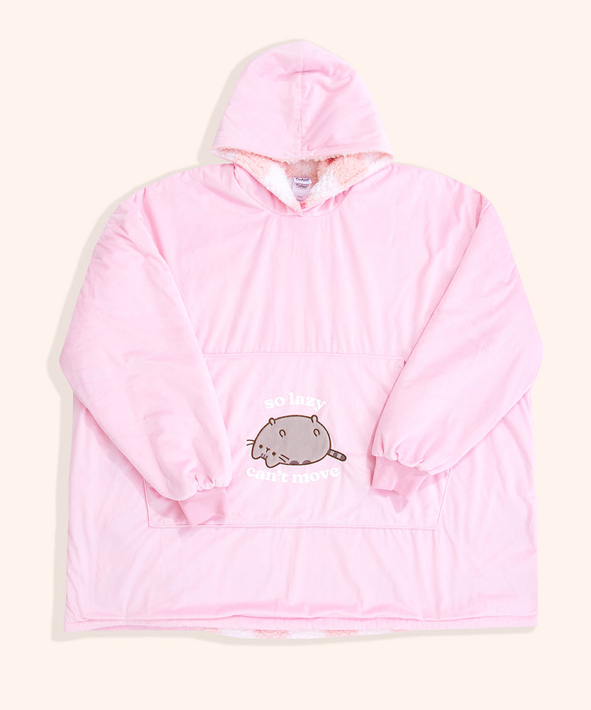 Pusheen Wearable Blanket Hoodie lies on a light yellow surface. The oversized pullover has a hood, oversized sleeves, a large front pocket, and an embroidered and flocked graphic of Pusheen the Cat. 