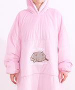 Front view of the model wearing the oversize blanket pullover sweater. The oversized sleeves are scrunched up to show the model’s hands. The large kangaroo style pocket sits towards the middle of the pullover. On the pocket is the flocked and embroidered Pusheen graphic where she’s laying and stating she’s “so lazy, can’t move.” 