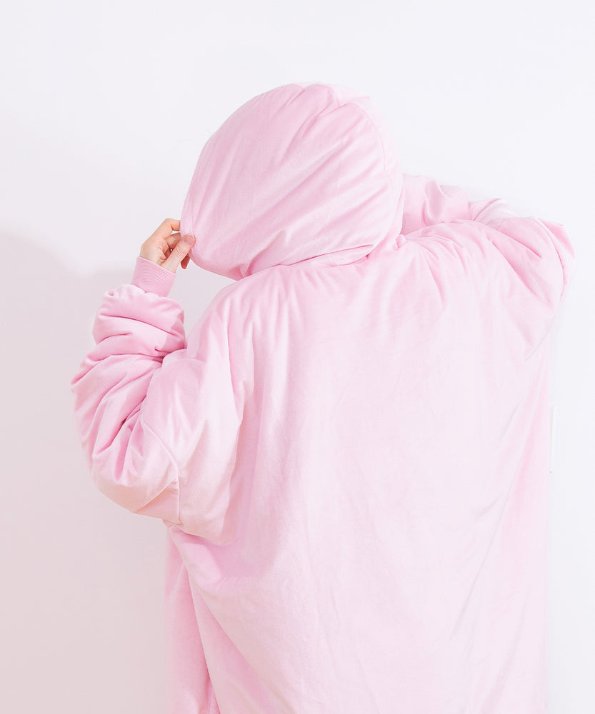 Back view of the model wearing the Wearable Blanket Hoodie. The light pink Pusheen pullover is oversized and the model holds the hood to the side to show the blanket hoodie’s size. 