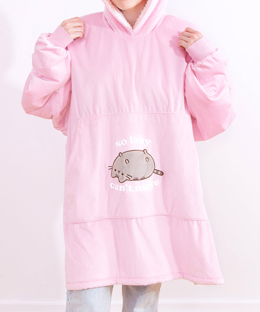 Model wears the Pusheen Wearable Blanket Hoodie paired with light denimjeans. Model is wearing the pink hood over her head, holding the pullover sweaters shoulders with both hands. The light pink hoodie is oversized and fits most. The hem of the sherpa-lined hoodie falls to the model’s knees. 