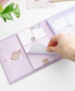 Model holds one sheet of paper in the included notepad. The notepad is pink and features a graphic of Pusheen the Cat holding a pink glazed donut at the bottom of the notepad. 
