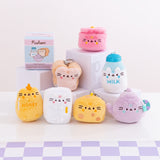 Front view of Pusheen’s Kitchen Surprise Plush assortment. Plush keychains sit on multi-level white pedestal in front of a white background with a purple checkered tabletop. Keychains feature various kitchen essentials including a pot, white bread slice, milk jug, honey pot, mini refrigerator, cheese slice, and tea pot. Accompanying the seven kitchen keychains is the mystery box packaging for the Kitchen Surprise Plush. 