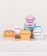 Front view of all Pusheen's Kitchen Surprise Squishy including the blind box capsule. The Squishys include a purple tea pot, white refrigerator, stack of pancakes, bread slice, and white milk bottle.