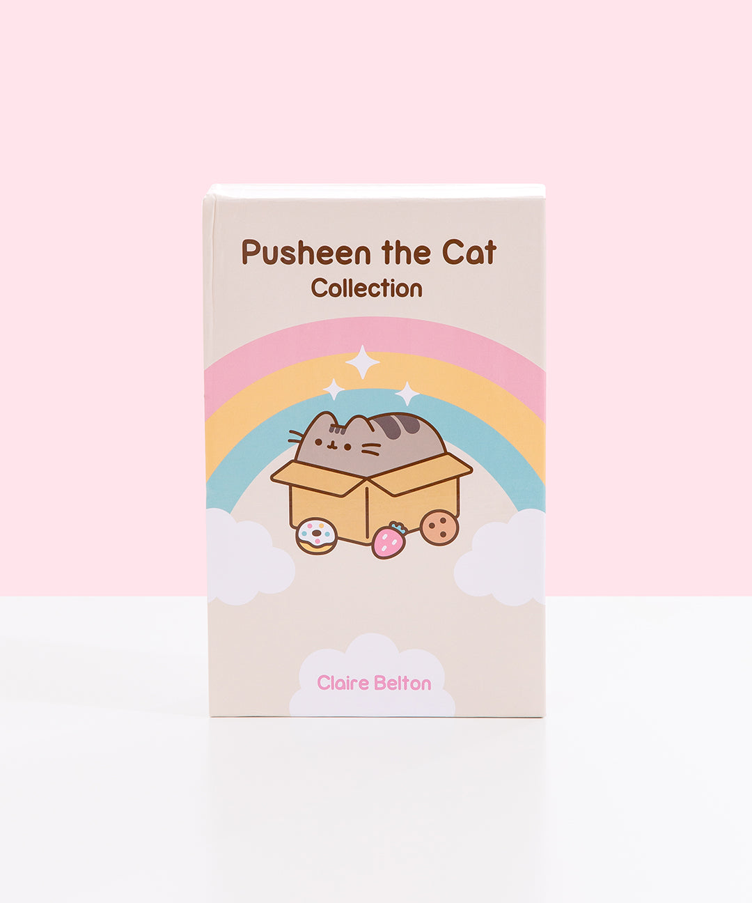 Front view of the Pusheen the Cat Collection: 3-Book Boxed Set. The decorative exterior storage box features the title of the boxed set. Below the title is a graphic of Pusheen laying inside a cardboard box while surrounded by a donut, strawberry, and chocolate chip cookie. Around Pusheen is a pink, yellow, and blue rainbow and white fluffy clouds and sparkles. At the bottom of the box is text stating the author, Claire Belton. 