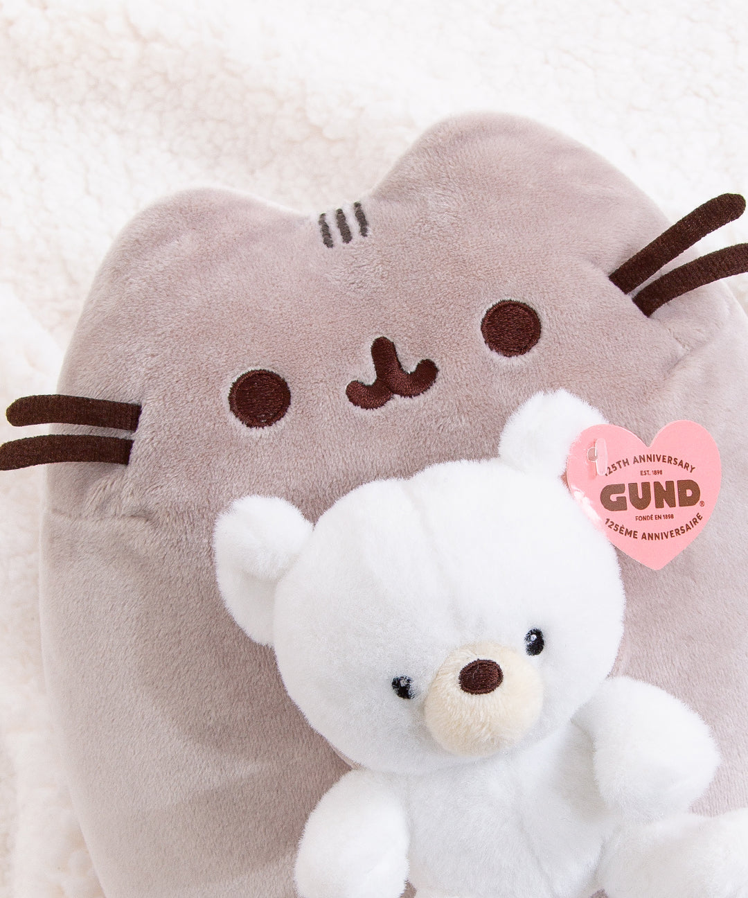 Close up view of the 2-in-1 plush featuring a grey Pusheen the Cat holding a white Kai bear from GUND. Pusheen's facial features are embroidered in brown and dark grey. The white anniversary plush bear features a cream colored snout and brown nose.