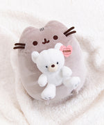 Pusheen x GUND Kai Bear Plush lies on a white fluffy surface. The grey cat holds a white teddy bear in front of her body.