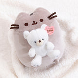Pusheen x GUND Kai Bear Plush lies on a white fluffy surface. The grey cat holds a white teddy bear in front of her body.