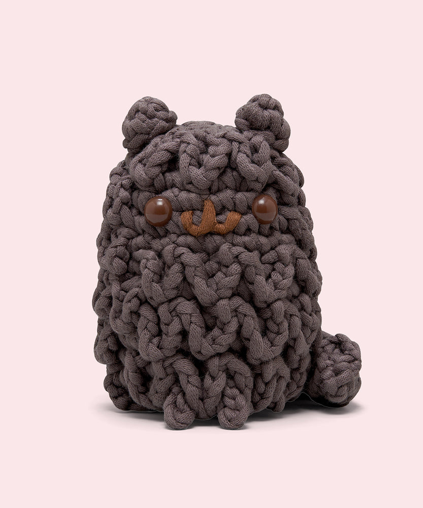 Front view of the Pusheen x The Woobles Pip Crochet Kit. The dark grey cat is crafted with yarn that mimics Pip’s unique shaggy fur pattern. The plush stands on two legs and has a tail feature. 