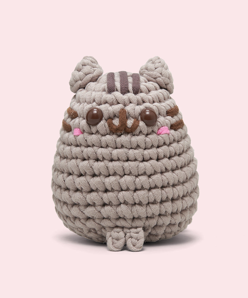 Front view of the Pusheen x The Woobles Pusheen Crochet Kit. The grey cat is crafted with yarn and features Pusheen's classic brown features including her head stripes, whiskers, smile, and eyes. A little pink blush is near the eyes of the plush.