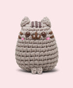 Front view of the Pusheen x The Woobles Pusheen Crochet Kit. The grey cat is crafted with yarn and features Pusheen's classic brown features including her head stripes, whiskers, smile, and eyes. A little pink blush is near the eyes of the plush.