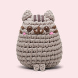 Front view of the Pusheen x The Woobles Pusheen Crochet Kit. The grey cat is crafted with yarn and features Pusheen's classic brown features including her head stripes, whiskers, smile, and eyes. A little pink blush is near the eyes of the plush.
