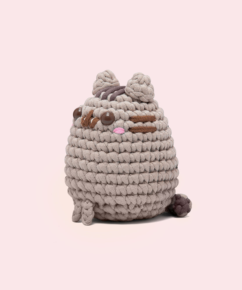 Side view of the Pusheen x The Woobles Pusheen Crochet Kit. The grey cat is crafted with yarn and features Pusheen's classic brown features including her head stripes, whiskers, smile, and eyes. A little pink blush is near the eyes of the plush. From this view, the crochet tail can be seen coming out of the bottom back of the figure. 