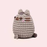 Side view of the Pusheen x The Woobles Pusheen Crochet Kit. The grey cat is crafted with yarn and features Pusheen's classic brown features including her head stripes, whiskers, smile, and eyes. A little pink blush is near the eyes of the plush. From this view, the crochet tail can be seen coming out of the bottom back of the figure. 