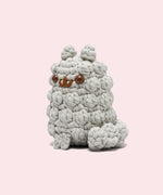 Side view of the Pusheen x The Woobles Stormy Crochet Kit. The light gray cat is crafted with yarn that mimics Stormy’s fluffy fur and tail. The plush stands on two legs and has a tail feature. From this view, the crochet tail can be seen coming out of the bottom back of the figure.