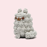 Side view of the Pusheen x The Woobles Stormy Crochet Kit. The light gray cat is crafted with yarn that mimics Stormy’s fluffy fur and tail. The plush stands on two legs and has a tail feature. From this view, the crochet tail can be seen coming out of the bottom back of the figure.