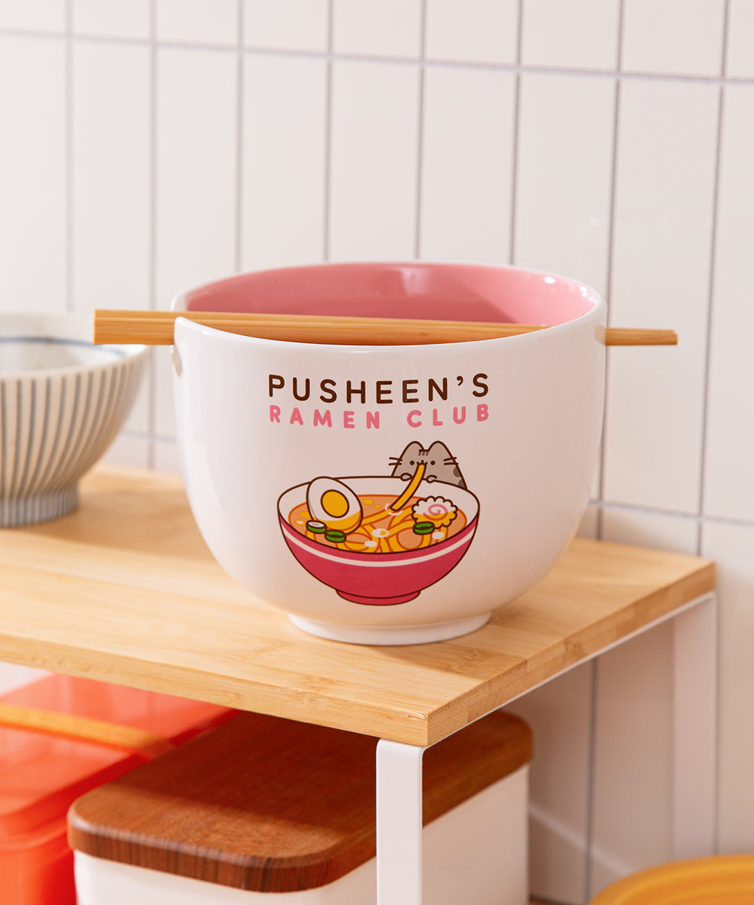Pusheen's Ramen Club Bowl sits on a counter with a pair of chopsticks resting in the dedicated slot at the rim of the bowl. The white bowl has a pink interior. 