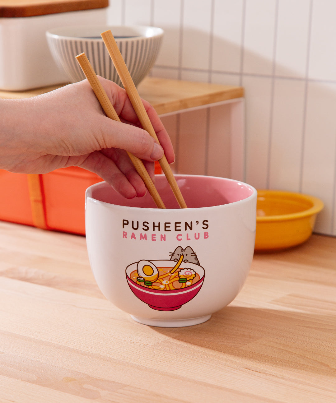 Model holds included wooden chopsticks above the white ceramic bowl that features a graphic of Pusheen enjoy her own bowl of ramen noodles.