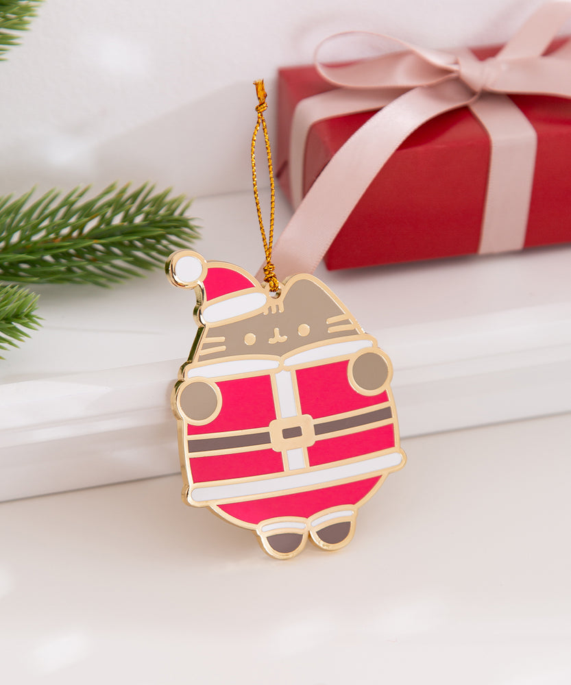 Pusheen Holiday Ornament rests on a white ledge and has a light shown on it to highlight the reflective gold outline details on the ornament. 