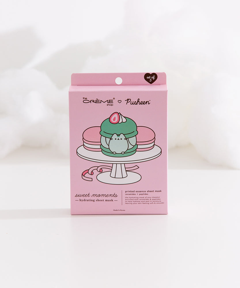 Pink sheet mask packaging box lies on white fluff. The packaging box shows graphics of three macarons on a dessert stand.