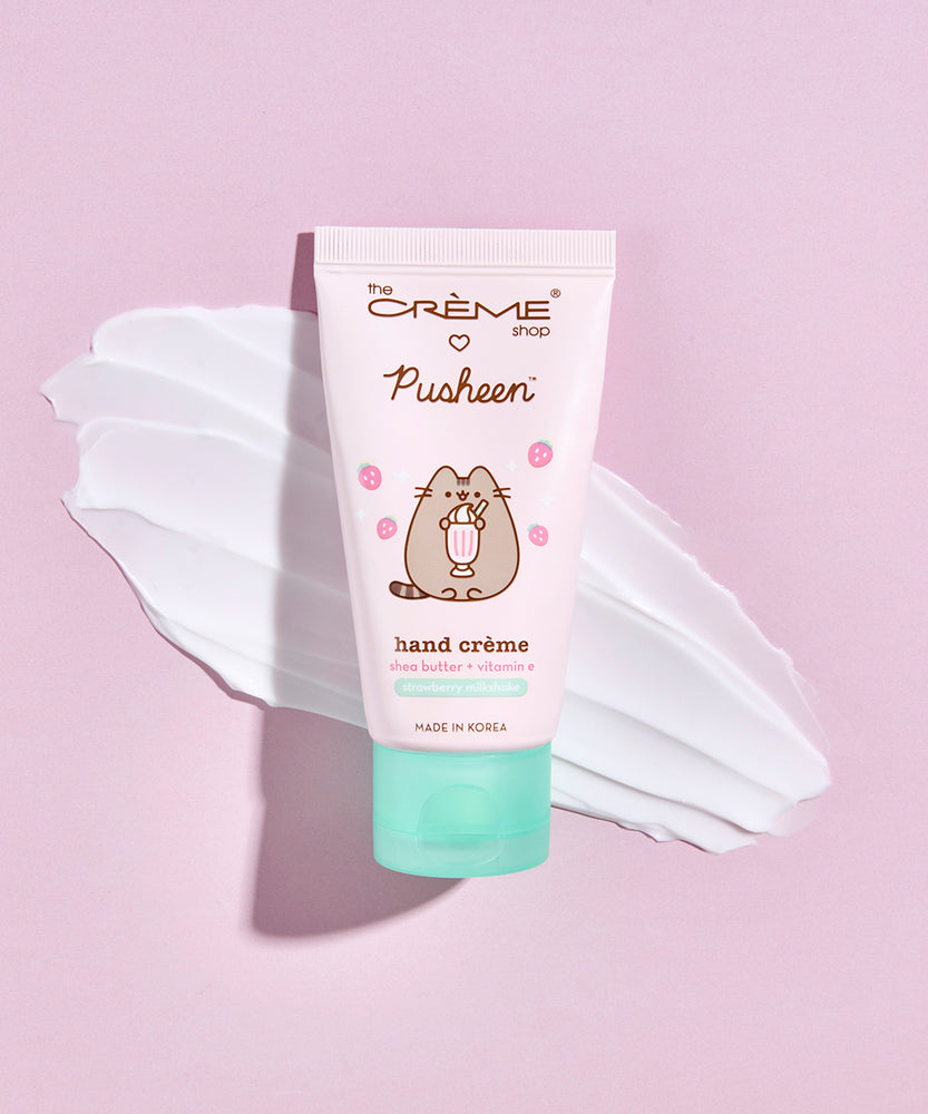 Front view of the Pusheen Strawberry Milkshake Hand Creme. The light pink tube has a printed graphic of Pusheen holding a milkshake surrounded by light pink strawberries. Behind the squeeze tube is a swatch of the white hand cream.