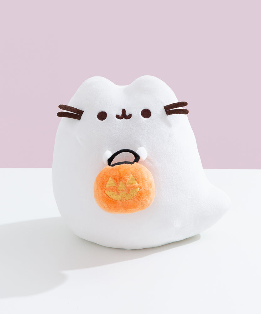 Front view of the white ghost plush that holds an orange candy pail that has yellow jack-o'-lantern details. The pail has two larger triangular eyes, a triangle nose, and a crooked carved mouth.