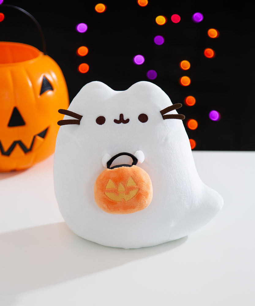 Front view of the Trick-or-Treat Boosheen Light Up Plush. The plush depicts Pusheen the Cat as a friendly ghost, Boosheen, and she holds a jack-o'-lantern candy pail.