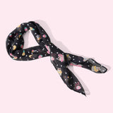 Pusheen Tropical Fruit Scarf lies on a light pink surface. The black chiffon-style scarf features an all over print of white polka dots, yellow pineapples, pink strawberries, and Pusheen and Stormy wearing inflatable pool floats.