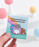 A close-up of the Pusheen Surprise Plush packaging. The front of the packaging features Pusheen as an enchanted raccoon in front of log stump, acorn, mushroom, and rainbow. The packaging says that these Surprise Plush are mini plush. 