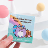 A close-up of the Pusheen Surprise Plush packaging. The front of the packaging features Pusheen as an enchanted raccoon in front of log stump, acorn, mushroom, and rainbow. The packaging says that these Surprise Plush are mini plush. 