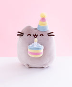 Pusheen sitting upright, eyes closed upwards and mouth open, with a striped party hat over her right ear. Her two nub paws hold a plush birthday cake with a single candle in it, and her bottom nub feet rest at the bottom. The plush is in front of a white and pink background.