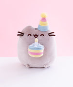 Pusheen sitting upright, eyes closed upwards and mouth open, with a striped party hat over her right ear. Her two nub paws hold a plush birthday cake with a single candle in it, and her bottom nub feet rest at the bottom. The plush is in front of a white and pink background.