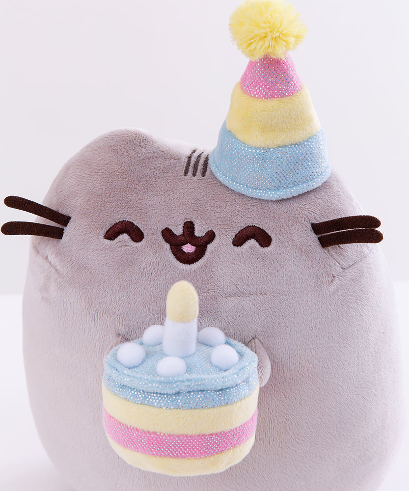 Close up of the Birthday Pusheen Plush. The party hat is composed of three stripes, a sparkly blue, a plush yellow, and a sparkly pink stripe, with a fuzzy yellow pompom on top. The plush cake Pusheen is holding is yellow with a sparkly pink filling, topped with sparkly blue icing, dots of white plush cream, and a plush candle in the middle.