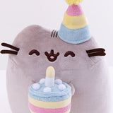 Close up of the Birthday Pusheen Plush. The party hat is composed of three stripes, a sparkly blue, a plush yellow, and a sparkly pink stripe, with a fuzzy yellow pompom on top. The plush cake Pusheen is holding is yellow with a sparkly pink filling, topped with sparkly blue icing, dots of white plush cream, and a plush candle in the middle.