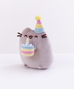 Quarter profile view of the Birthday Pushen Plush facing the left in front of a white background. Pusheen almost appears to be leaning forwards with her cake.