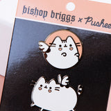 Close up of the Bishop Briggs x Pusheen Pin Set in it’s packaging. The Packaging is a rounded square, with a black square in the middle, orange rectangles on the top and bottom, and thin white lines in between. The Bishop Brigg x Pusheen Logo is printed on the top banner in black.