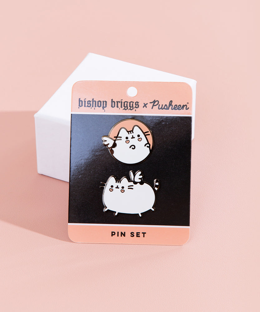 The Bishop Brigg x Pusheen Pin set in it’s original packaging leaning against a small square white pedestal, on top of a light orange background. The set includes two pins, one featuring a light orange circle with a white Pusheen with heart cheek marks and white wings inside, the other featuring a white Pusheen with black stripes galloping, pink heart marks underneath her eyes and two wings on top of her back that look like a butterfly.