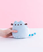 A model’s hand holding the Blue Mini Squisheen Plush in front of a white and pink background. The blue plush has dark blue stripes.