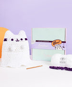 Crochet Boosheen plush posed next to the crochet kit box. The crochet hook, and fluffy white and purple yarn are displayed next to the box.