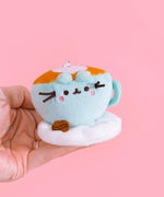 Close-up shot of Pusheen Breakfast character: coffee latte. Plush keychain is shaped like a coffee cup. Mint coffee cup sits on a white plush saucer with a brown coffee bean. The top of plush keychain is light brown with an inlaid white heart. 