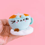 Close-up shot of Pusheen Breakfast character: coffee latte. Plush keychain is shaped like a coffee cup. Mint coffee cup sits on a white plush saucer with a brown coffee bean. The top of plush keychain is light brown with an inlaid white heart. 