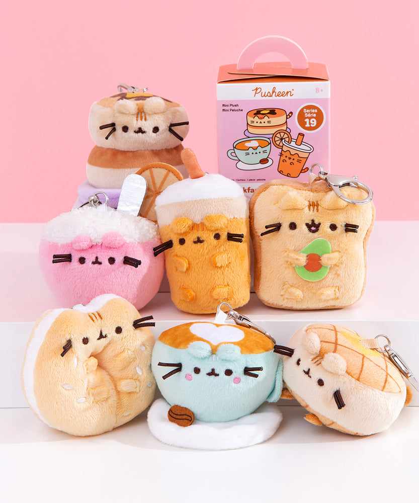 Front view of Pusheen Breakfast Surprise Plush assortment. Plush keychains sit on multi-level white pedestal in front of a pink background. Keychains feature food items including a pancake stack, cereal bowl, glass of orange juice, avocado toast, sesame bagel, coffee latte, and waffle. Accompanying the seven characters of breakfast Pusheens is the mystery box packaging for the Pusheen Breakfast Surprise Plush
